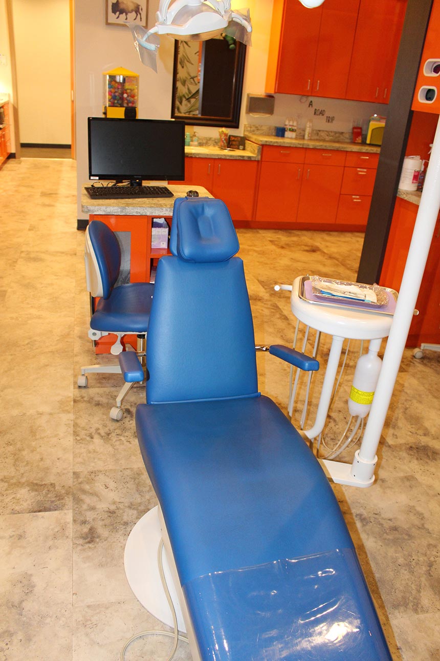 This is an interior view of a dental office with a blue dental chair and a countertop with various equipment and items, including a computer monitor.