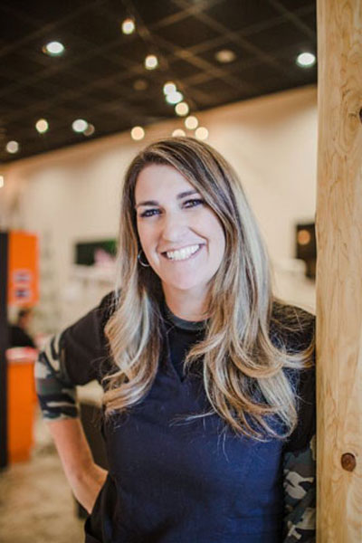 The image features a woman standing indoors with a friendly smile, wearing a dark top and leaning against a wall with a sign on it.