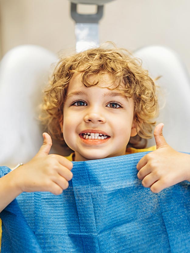 The image features a young child with curly hair giving a thumbs-up sign while wearing a blue surgical mask, seated in an examination chair, likely at a dentist s office.