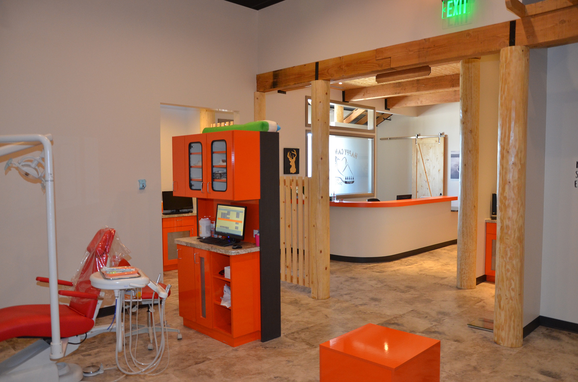 This is a photograph of an interior space resembling a salon or barber shop, with a modern design featuring orange and black accents on white walls, a wooden beam ceiling, a reception desk area with a computer monitor and a chair, and a large mirror reflecting the room's interior.