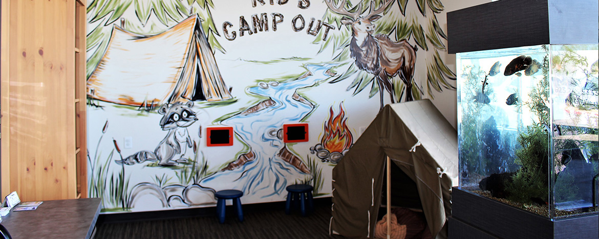 The image shows an indoor setting with a wall featuring a mural depicting a camping scene, and there's a person standing in front of the mural.