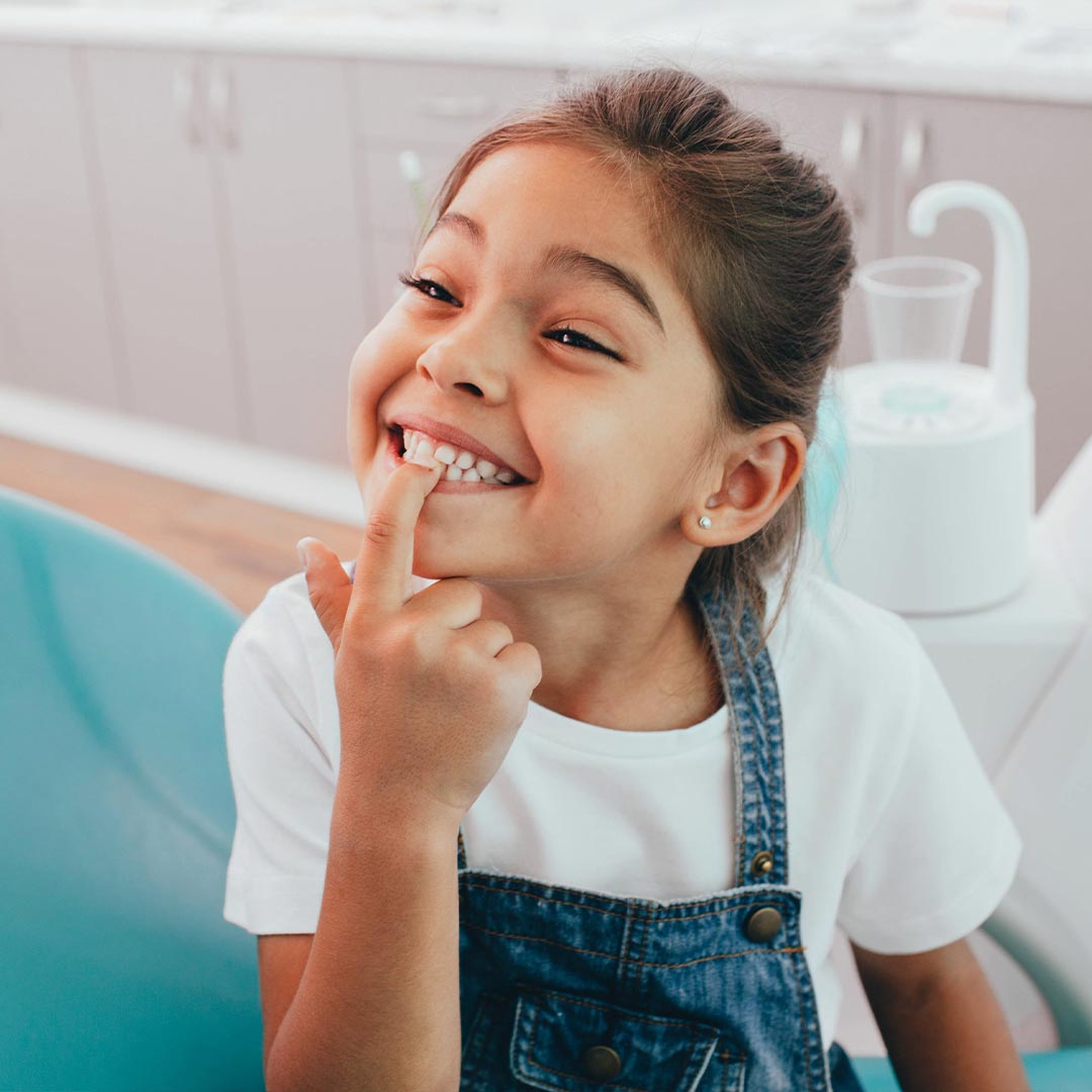 A young girl with her hand on her chin, smiling at the camera, sitting in a dental chair.