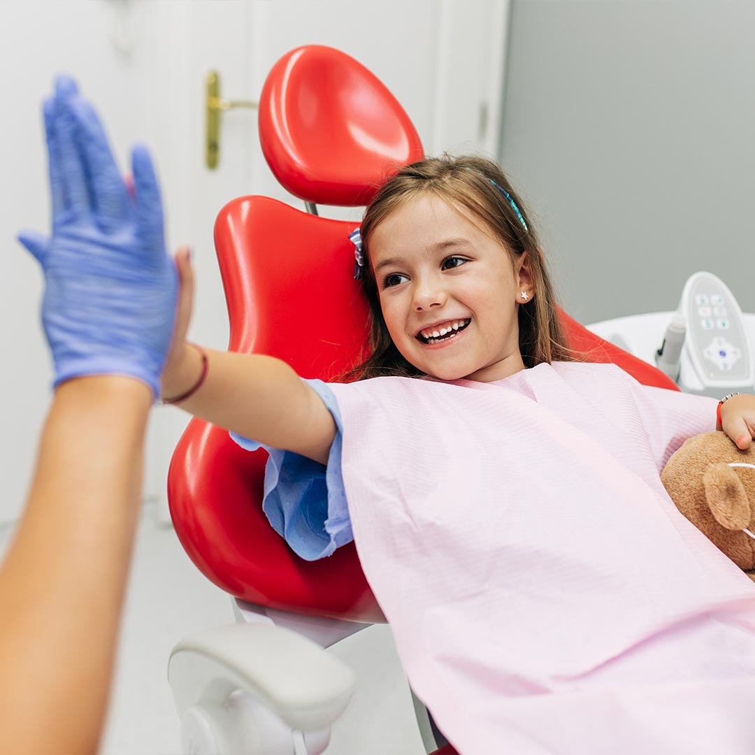 A young girl in a dental chair waving at someone with a smile, wearing blue gloves on her hands.