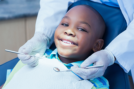 A young boy is sitting in a dental chair with a smile on his face, receiving dental care with a dentist working on him.